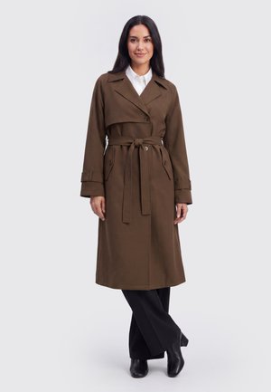 Donna in piedi che indossa un trench marrone con cintura sopra una camicia bianca, pantaloni neri e stivaletti neri su uno sfondo chiaro e uniforme.
