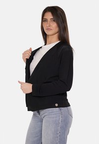 Cardigan nero a maniche lunghe con apertura frontale, tessuto texturizzato e un bottone decorativo all'orlo. Abbinato a una t-shirt bianca e jeans blu.