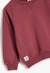 Sudadera de manga larga color burdeos con puños y cintura acanalados, pequeña etiqueta blanca cuadrada con una cara sonriente cerca del dobladillo inferior.