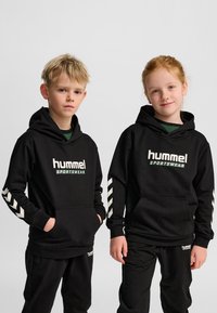 Sudaderas negras con capucha con el logo "hummel"; cuentan con detalles de chevrones blancos en las mangas; combinadas con pantalones negros a juego.
