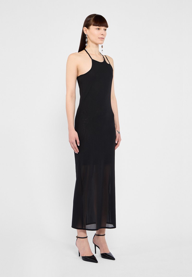 Femme debout dans une robe maxi noire sans manches avec un col halter et des talons hauts noirs sur un fond gris uni.
