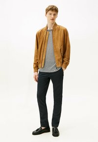 Giubbotto bomber in suede marrone sopra una camicia a righe bianche e nere, abbinato a pantaloni scuri e mocassini neri. Design semplice e privo di decorazioni.