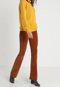 Un pull-over en tricot jaune à col roulé avec des manches longues, associé à un pantalon en velours côtelé marron à pattes d'éléphant, et des bottines noires brillantes.