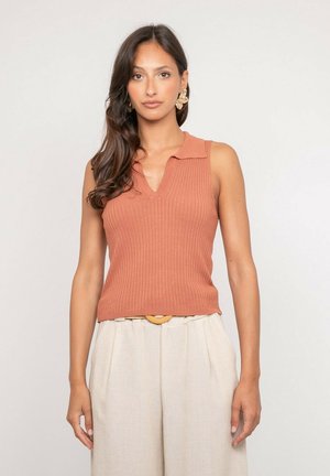 Femme aux longs cheveux foncés portant un haut sans manches côtelé de couleur rouille et un pantalon beige taille haute avec une ceinture, debout devant un fond uni.