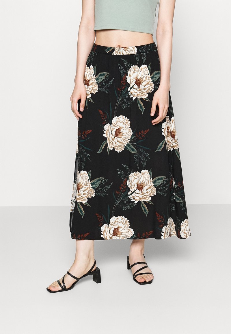 Falda maxi negra con flores grandes de peonías beiges y hojas verdes, con cintura elástica y aberturas laterales. Combinada con tacones negros de tiras.