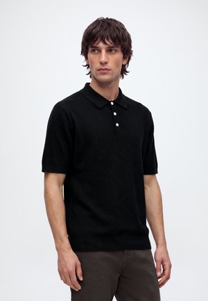 Jeune homme aux cheveux bruns mi-longs portant un polo noir à manches courtes et un pantalon foncé, debout devant un fond blanc uni.