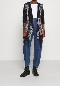 Svart kofta med vitt blomstertryck, öppen front, långa ärmar och fransdetaljer, kombinerad med högmidjade blå jeans och svarta stövlar.