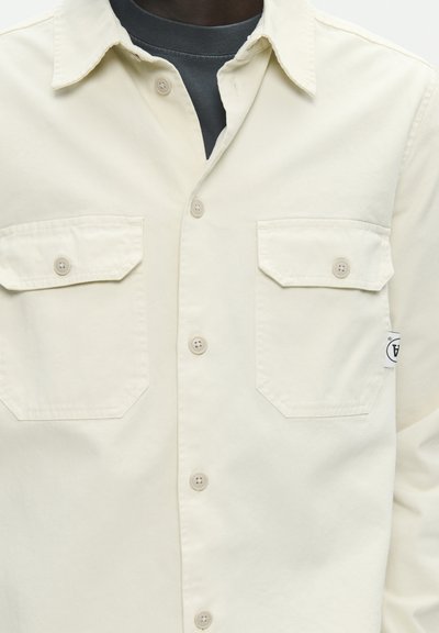 Homme portant une chemise crème à boutons avec deux poches poitrine par-dessus un t-shirt gris foncé à col rond, avec une petite étiquette blanche sur le côté droit.