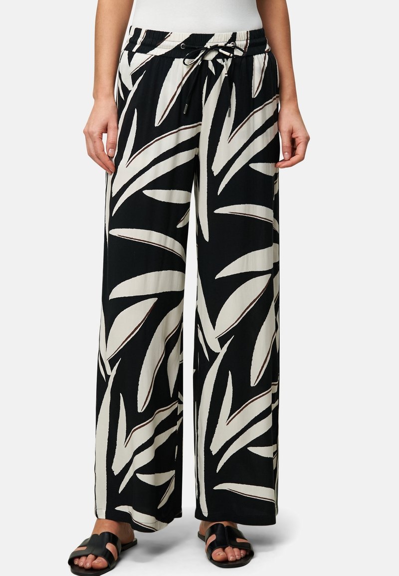 Femme portant un pantalon noir à jambes larges avec de grands motifs de feuilles blanches et des sandales noires à bout ouvert, se tenant avec les mains détendues sur les côtés.