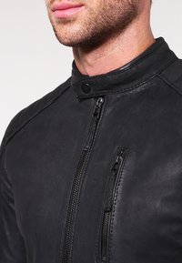 Giacca di pelle nera con colletto alto, dotata di chiusura frontale con zip e tasche con zip aggiuntive sul petto per lo stoccaggio.