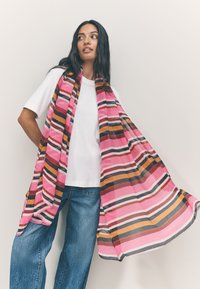 Sciarpa a righe dai toni rosa, arancione e marrone; materiale leggero con un design fluido; abbinata a una t-shirt bianca e jeans in denim.