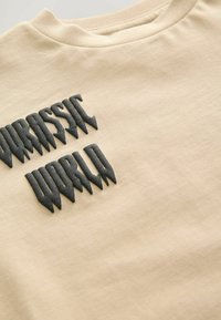 Beige bomulds t-shirt med sort, takkede "Jurassic World"-tekst i en hævet, struktureret design på forsiden.