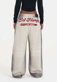 Pantalones de denim de pierna ancha en gris claro con un atrevido bordado rojo que dice "Ed Hardy" y "1971" en la parte posterior, con dos bolsillos traseros.