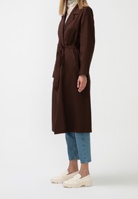Kvinna iklädd en mörkbrun lång kappa med bälte, ljusblå cropped jeans och krämfärgade loafers, stående mot en vit bakgrund.