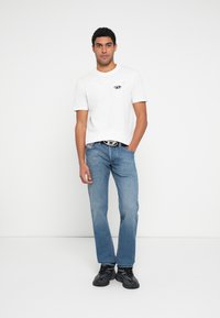 Λευκό κοντομάνικο μπλουζάκι με τυπωμένο λογότυπο, συνδυασμένο με ανοιχτό μπλε τζιν παντελόνι με ίσια γραμμή και μαύρα αθλητικά παπούτσια. Απλή, καθημερινή εμφάνιση.