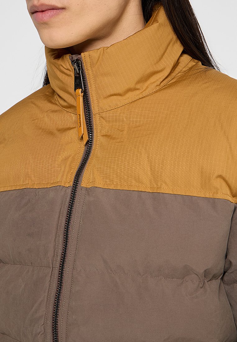 Piumino bicolore con parte superiore giallo oro e sezione inferiore marrone. Tessuto strutturato con collo alto e chiusura con cerniera.