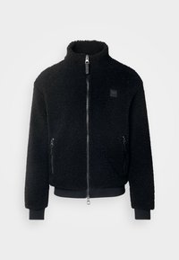 Giacca in pile nera con colletto alto, zip frontale, tasche con zip, polsini a coste e superficie testurizzata. Presenta una patch con logo sul petto.