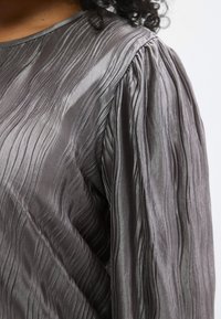 Szürke texturált blúz, gyűrött mintával és összegyűrt ujjakkal. Az anyag enyhén fényes és sima felületű.