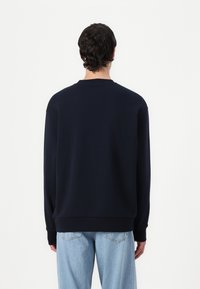 Felpa blu navy con maniche lunghe, collo a giro e polsini e orlo a coste. Tessuto morbido con una texture liscia. Indossata con jeans azzurri.