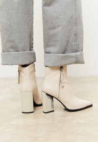 Beige keinilahko ankle boots dengan ujung runcing, detail ritsleting, dan hak blok perak bermotif geometris. Dipakai dengan kerah denim abu-abu yang digulung.