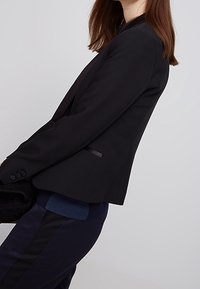 Svart skräddarsydd kavaj med slim fit, mjukt tyg, satinädelningar och två knappar, stylar med en lager av marinblå topp och mörka byxor.