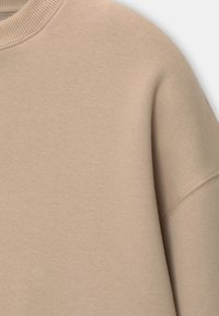 Détail du tissu sweat-shirt beige montrant un col ras du cou côtelé et une couture sur la manche longue, texture coton lisse.