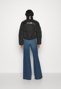 Giacca a vento nera con spalle lucide e lisce, design cropped e jeans in denim blu a gamba larga. Sfondo neutro e semplice.