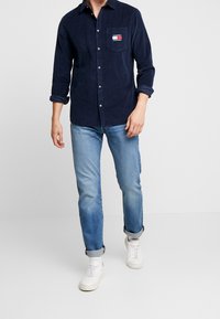 Marineblauwe langdriedige shirt met een button-down kraag en een borstzak. Lichtblauwe spijkerbroek en witte sneakers maken de outfit compleet.