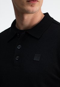 Schwarzes PoloheMD, gefertigt aus strukturiertem Stoff, mit einem Kragen, drei Knöpfen und einem kleinen Logo-Patch auf der Brust.