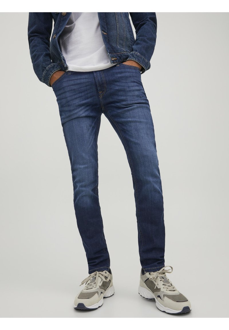 Pantalones vaqueros de mezclilla en azul oscuro, corte ajustado, con un sutil efecto desgastado. Combinados con zapatillas deportivas en gris y blanco, que presentan un diseño deportivo.