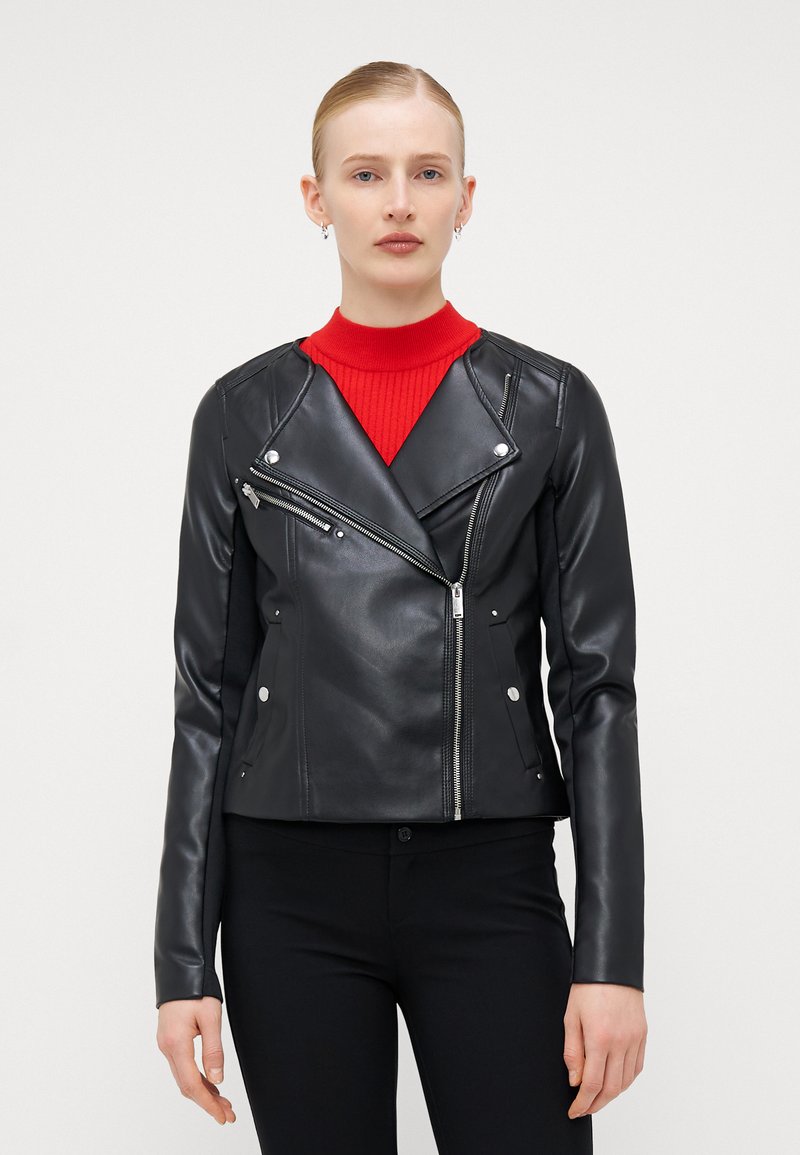 Veste de motard en faux cuir noir avec une fermeture éclair asymétrique à l'avant, quincaillerie argentée et poches zippées, associée à un pull rouge à col montant.