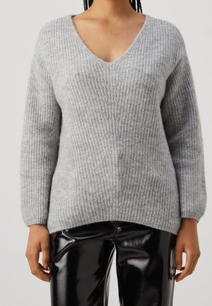 Femme portant un pull gris à côtes avec col en V et un pantalon noir en vinyle brillant, debout devant un fond clair uni.