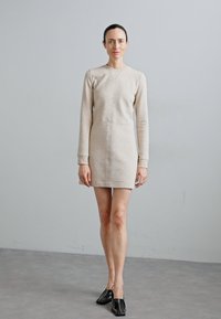 Beige kleit-sweatshirt pikade varrukatega, ümar kaelus ja keha lähedane lõige. Omab keskmist õmblust ja ribakudumist randmetes, koos mustade kingadega.