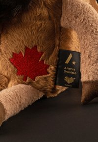Plush toy featuring brown and beige fur, a red embroidered maple leaf, and a black label with gold text reading "America Unlimited Canada Kollektion."