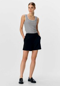 Zwarte shorts met zijzakken, gecombineerd met een mouwloos wit tanktop met dunne zwarte strepen. Model draagt glanzende zwarte instappers.