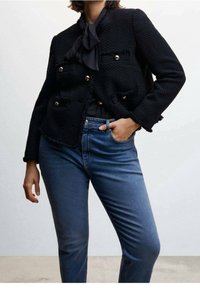 Chaqueta negra texturizada con botones dorados y bolsillos, combinada con jeans ajustados azules. La chaqueta presenta un diseño corto y bordes deshilachados.