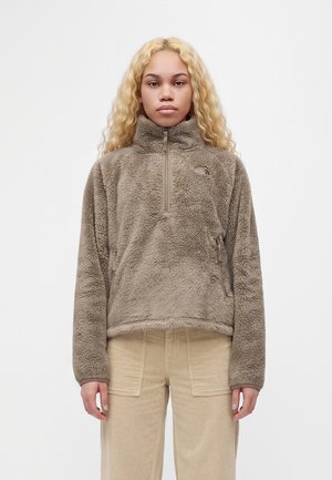 Donna con lunghi capelli biondi ricci che indossa una giacca in pile color taupe con zip e pantaloni beige strutturati, in piedi davanti a uno sfondo bianco semplice.