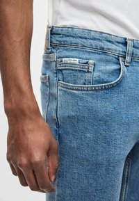 Svetlomodré džínsové nohavice s úzkym strihom, päťvreckovým dizajnom a malou značkovou nášivkou na zadnej strane, s viditeľnými švami.