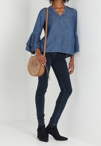 Top in denim con maniche a campana, jeans skinny scuri e stivali neri alla caviglia. Borsa a tracolla circolare color tan. Outfit casual con una silhouette rilassata.