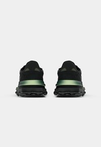 Scarpe da ginnastica nere con tessuto texturizzato e una fascia verde accentata, dotate di una suola nera robusta progettata per garantire trazione.