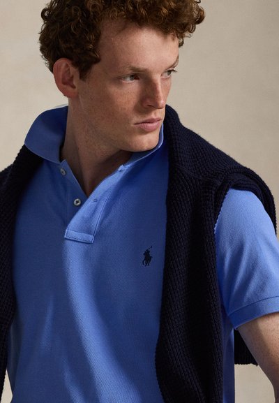 Jonge man met krullend haar die een blauw poloshirt en een donkergebreide trui over zijn schouders draagt, kijkt opzij.