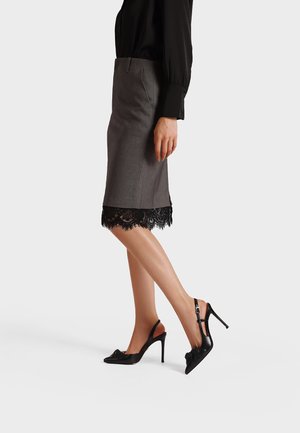 Donna che indossa una gonna a tubino grigia con rifiniture in pizzo nero, camicetta nera e scarpe nere con tacco alto e cinturino posteriore su sfondo bianco.