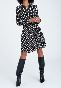 Abito camicia a pois neri e crema con colletto, chiusura a bottoni e vita regolabile, abbinato a stivali neri di pelle al ginocchio.