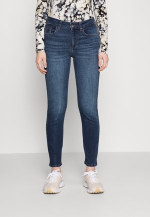 Femme portant un jean skinny bleu foncé, un haut à manches longues à motifs, et des baskets blanches, debout devant un fond clair uni.