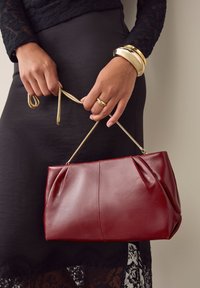 Rode leren handtas met gevouwen ontwerp en gouden kettinghengsel. Gladde textuur, rechthoekige vorm, gecombineerd met een zwarte kanten rok.