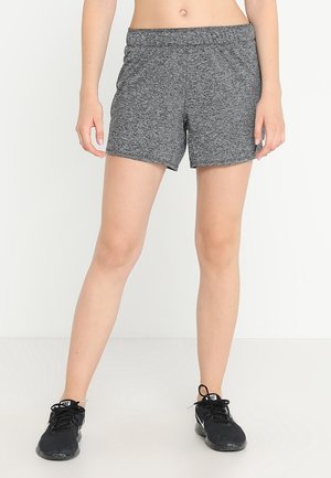 Frau trägt graue Sportshorts und schwarze Nike Laufschuhe, steht vor einem schlichten weißen Hintergrund.