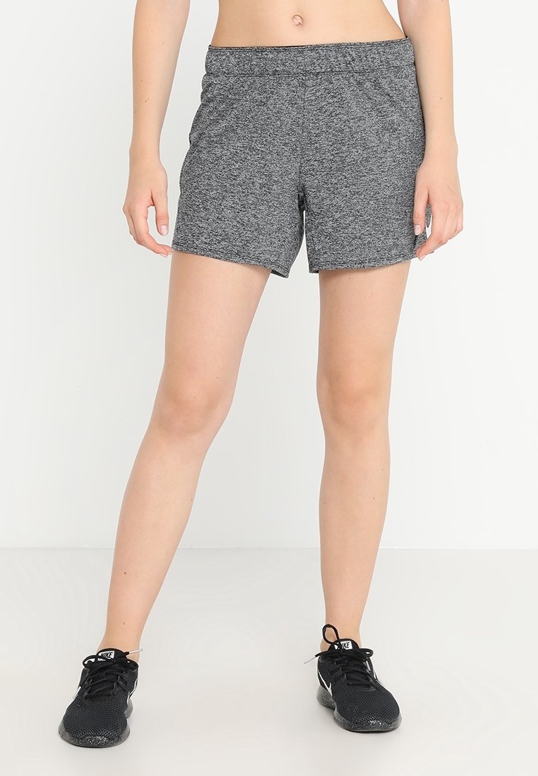 Femme portant un short de sport gris et des chaussures de course Nike noires, debout sur un fond blanc uni.
