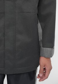 Grå jacka med en slät textur, med ett fiskbensmönster och en kontrasterande grå manschett. Inkluderar sidofickor och detaljer med dragkedja.