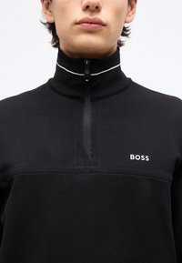 Jeune personne portant un pullover noir à demi-fermeture éclair avec un col montant et un logo blanc "BOSS" sur la poitrine, sur un fond uni.