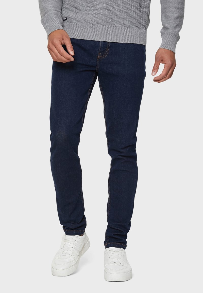 Jeans ajustados de color azul oscuro, fabricados en denim, con un diseño clásico de cinco bolsillos y costuras en contraste, combinados con zapatillas blancas y un suéter gris.